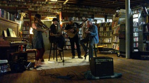 A small band performs in a cozy library filled with books, featuring a violinist, guitarist, and two musicians.
