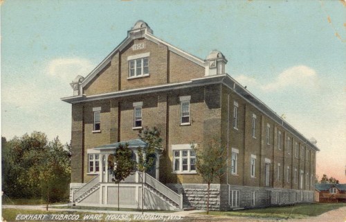 Historic brick building with a gabled roof, labeled "Eckhart Tobacco Warehouse," surrounded by trees.