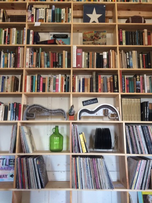 A wooden bookshelf filled with books and vinyl records, featuring decorative items and a Texas flag at the top.