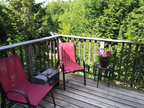 A wooden balcony with two red chairs, a small table, and a potted plant, surrounded by lush green trees.