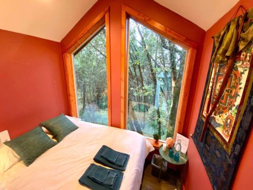 Cozy bedroom with a bed, green pillows, and large windows overlooking a forested area. Decorated with wall art.