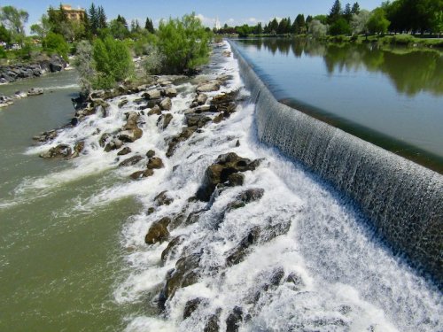 A serene river scene featuring a waterfall cascading over rocks, surrounded by lush greenery and clear blue skies.