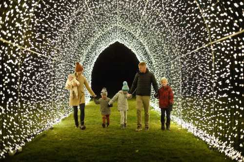 A family walks hand-in-hand through a sparkling light tunnel, surrounded by glowing decorations at night.