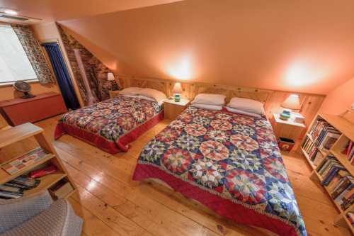 Cozy attic bedroom with two beds, colorful quilts, wooden walls, and warm lighting. Bookshelf in the corner.