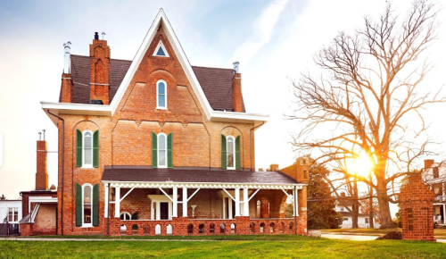 A large brick house with green shutters, featuring a porch, set against a sunset and a bare tree.