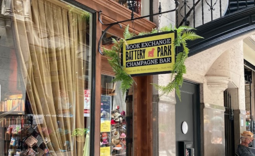 Sign for "Book Exchange Battery Park Champagne Bar" with greenery, located near a storefront with curtains.