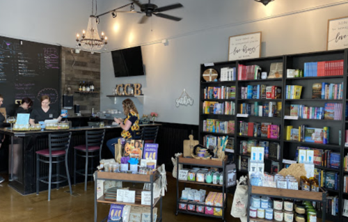 A cozy café interior with a bar, shelves of books, and various products for sale, featuring patrons at the counter.