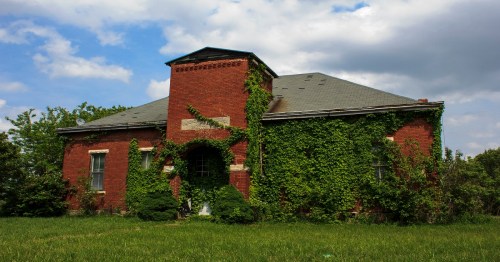 Abandoned brick building covered in green ivy, set against a cloudy sky and surrounded by overgrown grass.