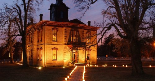 A historic brick house illuminated at dusk, surrounded by trees and lined with glowing candles along a pathway.