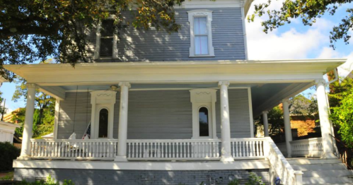 A large, two-story gray house with white trim, featuring a wraparound porch and tall columns under a blue sky.