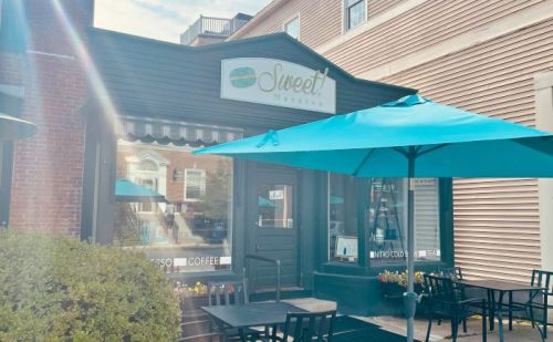 Exterior of a café named "Sweet!" with teal umbrellas and outdoor seating. Bright and inviting atmosphere.