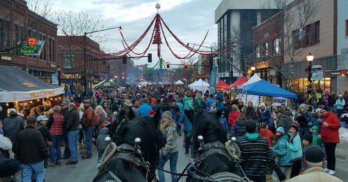 A bustling street festival with crowds, tents, and holiday decorations, viewed from behind two horses.