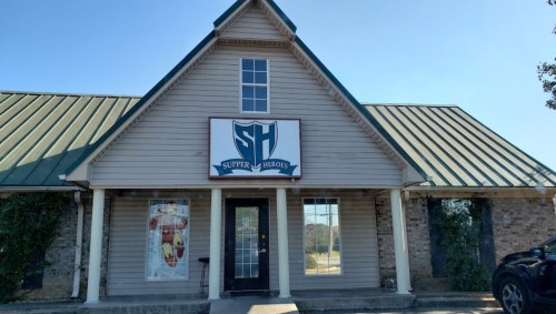 Exterior of a building with a sloped roof, featuring a sign that reads "Supper Heroes" prominently displayed.