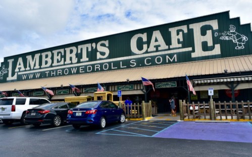 Exterior of Lambert's Cafe with a green sign, parked cars, and American flags outside.