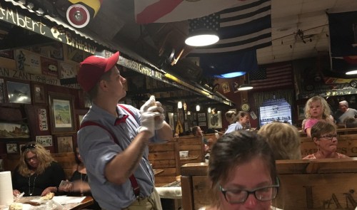 A waiter in a red cap and gloves serves food in a busy restaurant decorated with flags and vintage memorabilia.