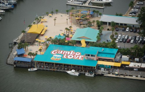 Aerial view of a colorful waterfront restaurant named "Gumbo Love" with a beach area and boats nearby.