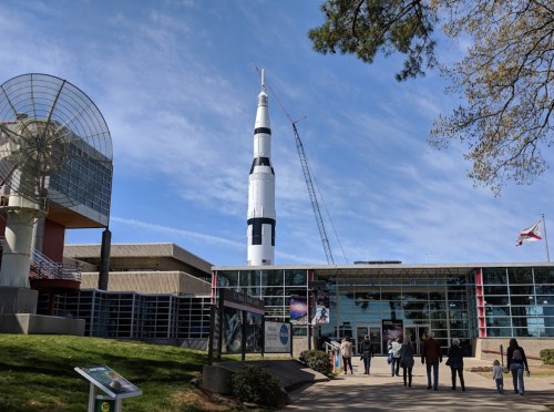 A large rocket stands outside a modern building, with visitors walking towards the entrance under a blue sky.