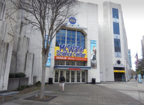 Exterior of the McWane Science Center with IMAX Dome Theater signage, featuring a tree and a person walking by.