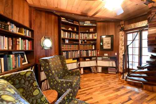 Cozy library with wooden walls, bookshelves filled with books, two patterned chairs, and a spiral staircase.