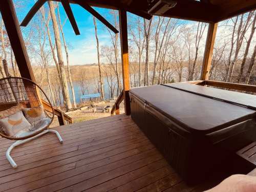 A wooden deck with a hot tub overlooking a serene lake surrounded by trees on a clear day.