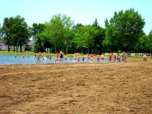 A sandy beach with children playing and wading in a shallow lake, surrounded by green trees and a clear blue sky.
