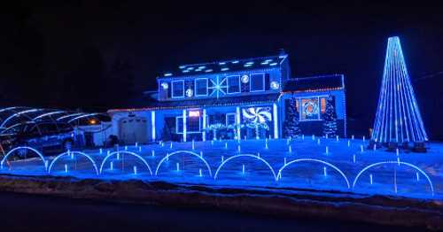 A house decorated with vibrant blue lights, featuring a tall lighted tree and illuminated pathways in the snow.