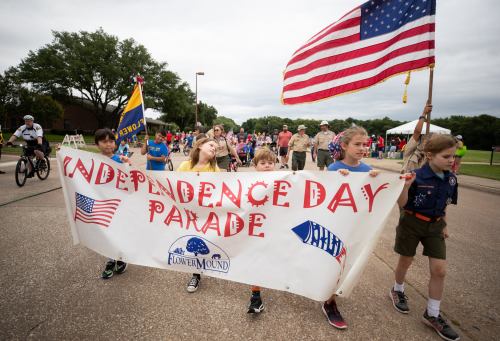 Children and adults march in a parade, holding a banner that reads "Independence Day Parade" with flags in the background.