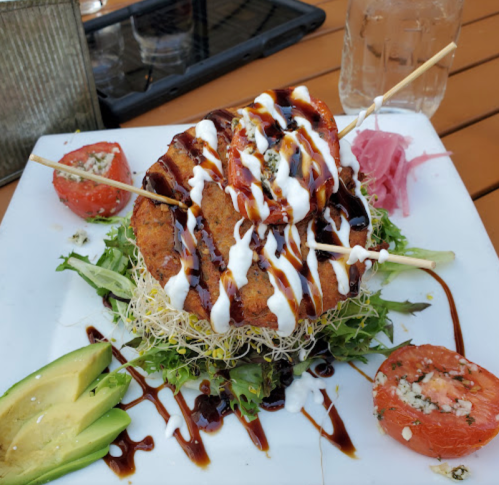 A plate of food featuring a fried patty topped with sauce, served on a bed of greens, avocado, and garnished with tomatoes.
