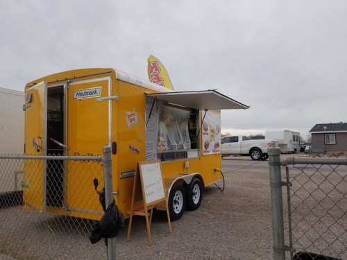 A bright yellow food truck with an open window, parked behind a chain-link fence on a cloudy day.