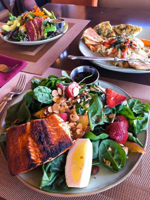 A plate of spinach salad topped with grilled salmon, strawberries, almonds, and a lemon wedge, with other dishes in the background.
