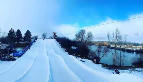 Snow-covered tubing hill with a blue sky, trees, and a river in the background.