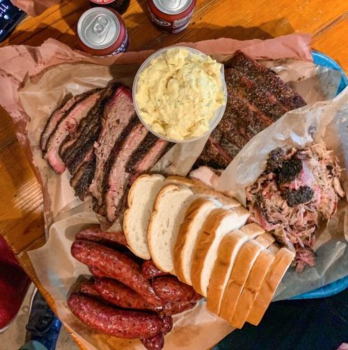 A platter of barbecue featuring brisket, pulled pork, sausages, bread, and a side of potato salad.