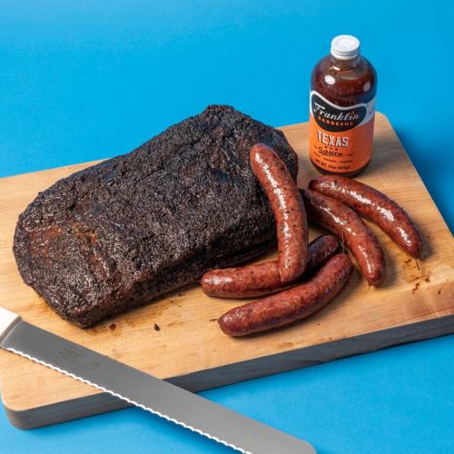 A wooden cutting board with smoked brisket, sausages, and a bottle of Texas barbecue sauce on a blue background.