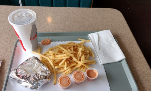 A tray with a soda, a serving of fries, a wrapped burger, and three dipping sauces on a table.