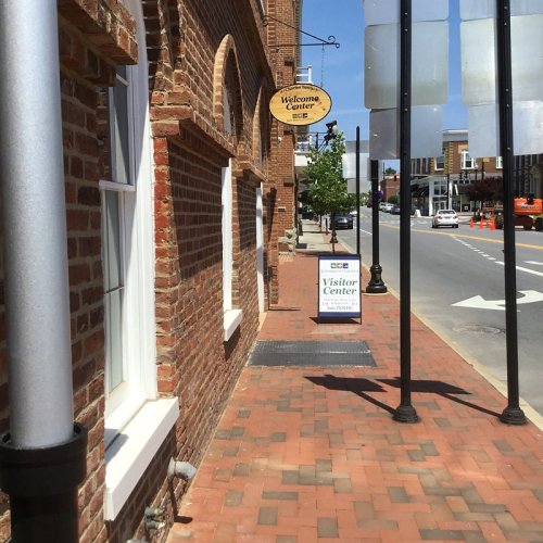 A brick building with a welcome sign and a visitor center sign on a sunny street with trees and parked cars.