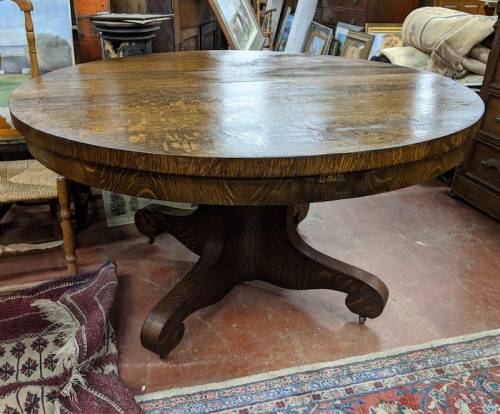 A round wooden table with a decorative surface and a sturdy pedestal base, set in an antique shop.
