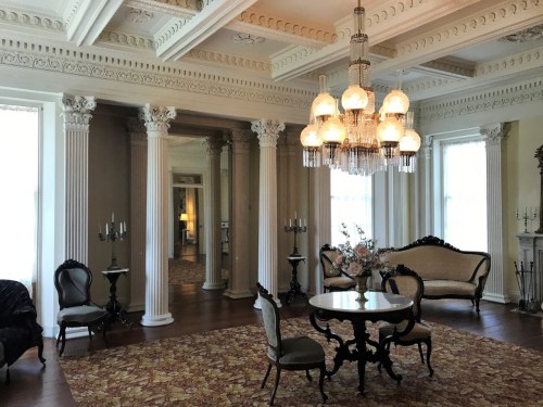 Elegant room with ornate columns, a chandelier, vintage furniture, and a decorative rug. Natural light fills the space.