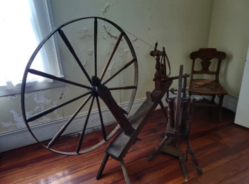 A vintage spinning wheel made of wood, positioned in a room with peeling paint and wooden floors.