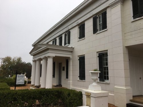 A white historic building with columns, black shutters, and a sign, surrounded by neatly trimmed hedges.
