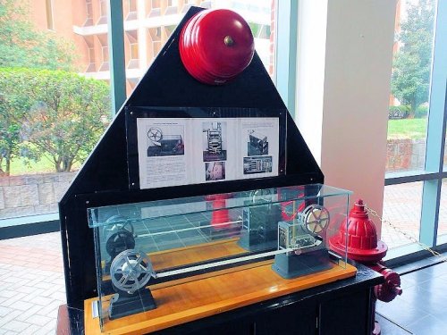 Display featuring a fire alarm system model, with a bell, glass case, and informational panels in a well-lit area.