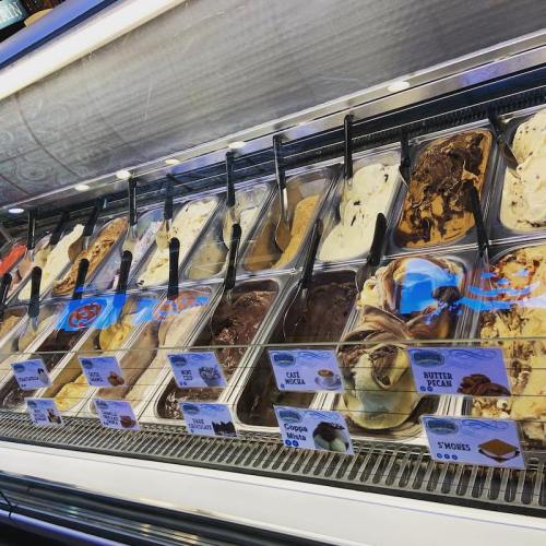 A display of various gelato flavors in metal containers, with scoops and labels for each flavor.