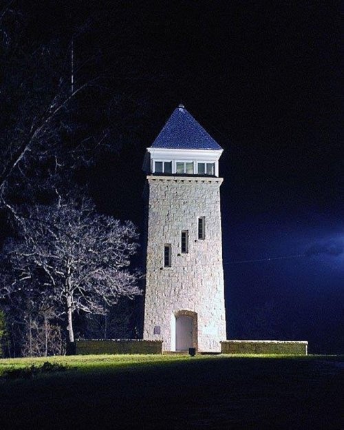 A tall stone tower with a blue roof, illuminated at night, surrounded by dark trees and a cloudy sky.