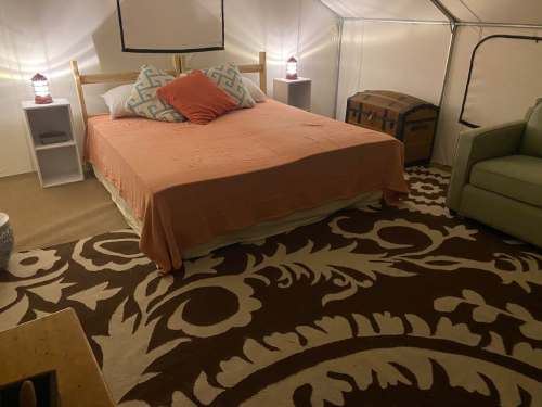 Cozy tent interior featuring a bed with orange bedding, decorative pillows, a rug, and a green chair.