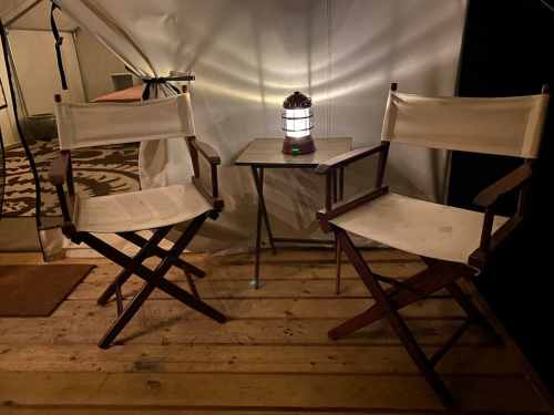 Two white canvas chairs beside a small table with a lantern, set in a cozy tent interior with wooden flooring.