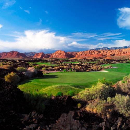 A scenic view of a golf course surrounded by red rock formations and blue skies, with lush green grass in the foreground.