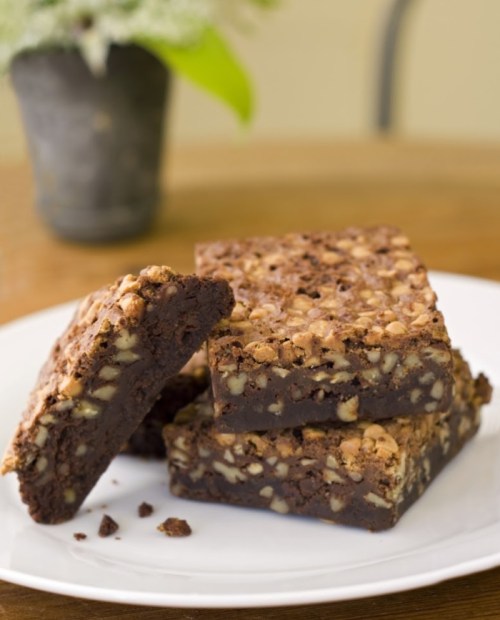 A plate of rich, nutty brownies stacked with one leaning against the others, set on a wooden table.