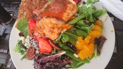 A colorful salad topped with grilled salmon, strawberries, oranges, and blue cheese, served on a white plate.