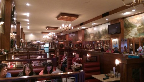 A cozy restaurant interior with wooden decor, chandeliers, and patrons enjoying meals at booths and tables.