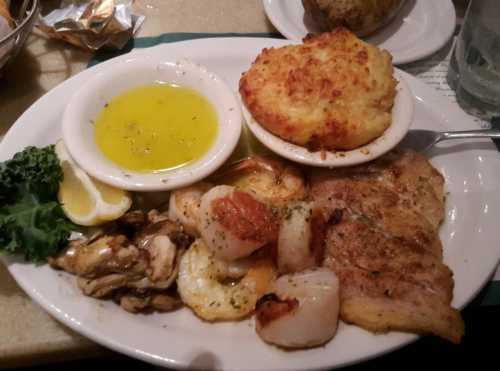 A plate of seafood including shrimp, scallops, and fish, served with a side of cornbread and lemon wedges.