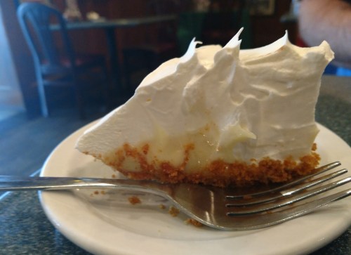 A slice of pie with a graham cracker crust, topped with fluffy whipped cream, on a plate with a fork beside it.